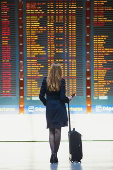 Young woman in international airport looking at the flight information board, holding passport in her hand, checking her flight