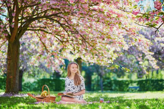 Beautiful young woman having picnic on sunny spring day in park during cherry blossom season, reading a book