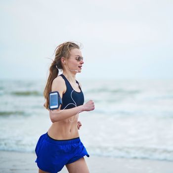 Running fitness woman wearing phone armband monitor tracker. Jogger running fast on beach and listening to music using earphones. Fitness and healthy lifestyle concept