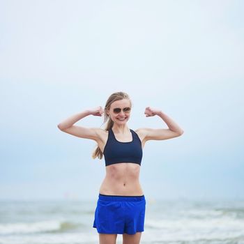 Healthy fitness runner girl wearing sunglasses showing her biceps. Young European woman on beach cardio training taking a rest during workout