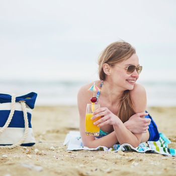 Beautiful young woman relaxing and sunbathing on beach, drinking delicious fruit or alcohol cocktail with paper umbrella, beach bag on sand near the model