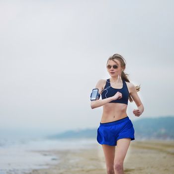 Running fitness woman wearing phone armband monitor tracker. Jogger running on beach and listening to music using earphones. Fitness and healthy lifestyle concept
