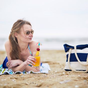 Beautiful young woman relaxing and sunbathing on beach, drinking delicious fruit or alcohol cocktail with paper umbrella, beach bag on sand near the model