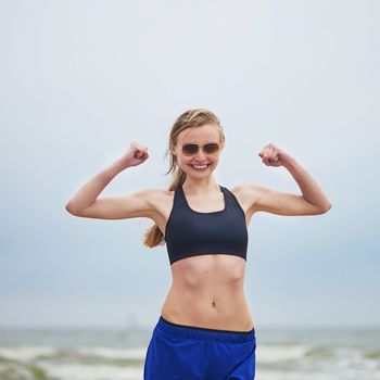 Healthy fitness runner girl wearing sunglasses showing her biceps. Young European woman on beach cardio training taking a rest during workout. Healthy lifestyle and fitness concept