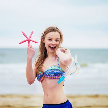Happy young woman in bikini with snorkelling equipment and pink starfish enjoying summer vacation holidays by ocean or sea. Beach, travelling and people concept