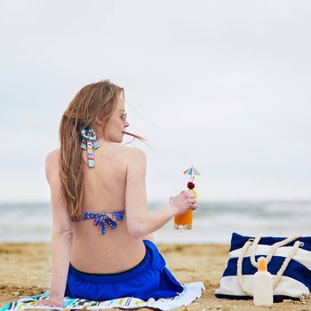 Beautiful young woman relaxing and sunbathing on beach, drinking delicious fruit or alcohol cocktail with paper umbrella, beach bag and sunscreen bottle on sand near the model