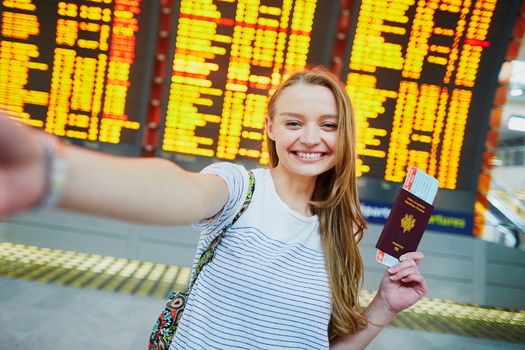 Beautiful young tourist girl in international airport, taking funny selfie with passport and boarding pass near flight information board