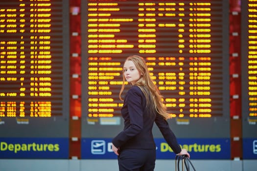 Young elegant business woman with hand luggage in international airport terminal, looking at information board, checking her flight. Cabin crew member with suitcase.