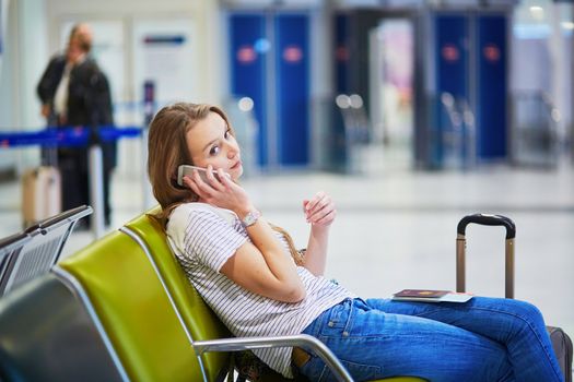 Young traveler with carry on luggage in international airport checking her mobile phone while waiting for her flight