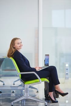 Young elegant business woman with hand luggage in international airport terminal, working on her laptop while waiting for flight