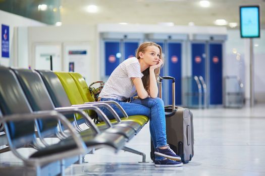 Beautiful young tourist girl with backpack and carry on luggage in international airport, waiting for her flight, looking upset. Delayed or canceled flight concept