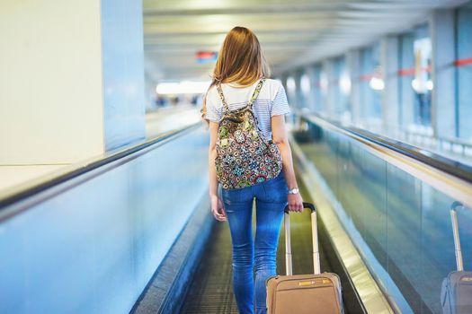 Beautiful young tourist girl with backpack and carry on luggage in international airport, on travelator