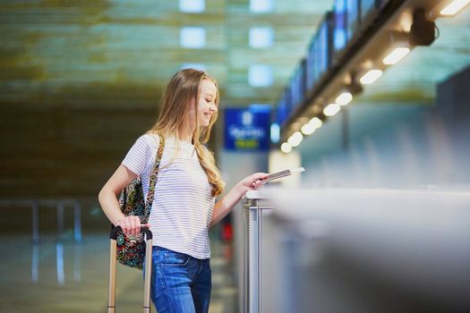 Beautiful young tourist girl with backpack and carry on luggage in international airport at check-in counter, giving her passport to an officer and waiting for her boarding pass