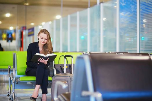 Young elegant business woman with hand luggage in international airport terminal, reading book while waiting for flight