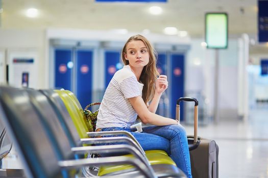 Beautiful young tourist girl with backpack and carry on luggage in international airport, waiting for her flight, looking upset. Delayed or canceled flight concept