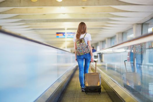 Beautiful young tourist girl with backpack and carry on luggage in international airport, on travelator