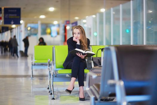 Young elegant business woman with hand luggage in international airport terminal, reading book while waiting for flight
