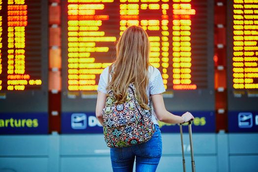 Beautiful young tourist girl with backpack and carry on luggage in international airport, near flight information board