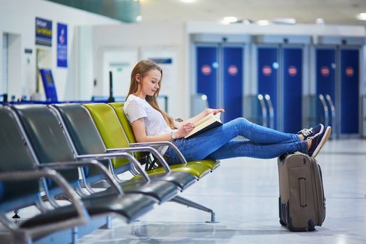 Beautiful young tourist girl with backpack and carry on luggage in international airport, reading a book while waiting for her flight