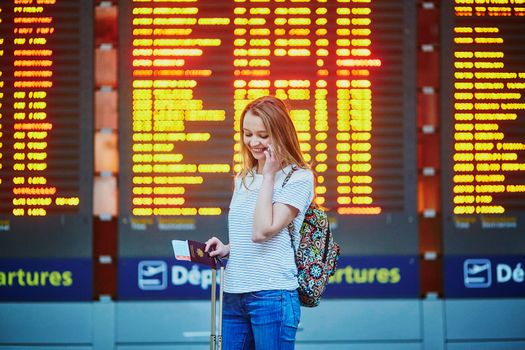 Beautiful young tourist girl with backpack and carry on luggage in international airport, near flight information board