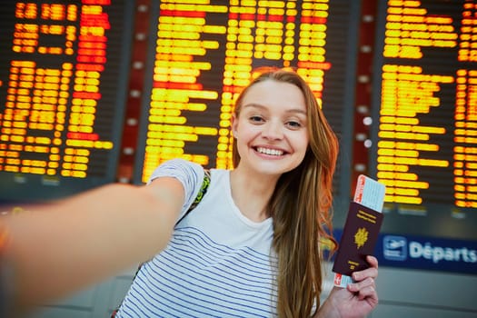 Beautiful young tourist girl in international airport, taking funny selfie with passport and boarding pass near flight information board