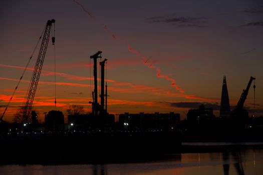 A sunset cityscape with The Shard skyscraper building and tower cranes silhouetted against an orange sky with aeroplane trails