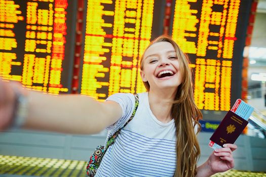 Beautiful young tourist girl in international airport, taking funny selfie with passport and boarding pass near flight information board