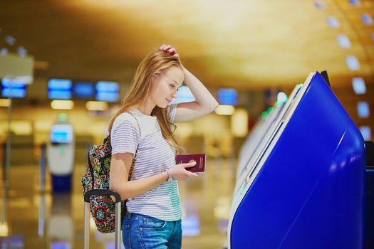 Young tourist girl with backpack and carry on luggage in international airport, doing self check-in, looking upset and worried. Delayed or missed flight concept