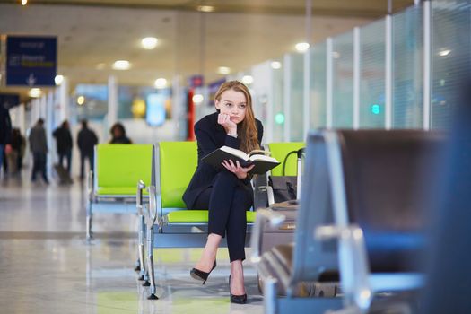 Young elegant business woman with hand luggage in international airport terminal, reading book while waiting for flight