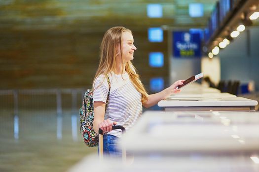 Beautiful young tourist girl with backpack and carry on luggage in international airport at check-in counter, giving her passport to an officer and waiting for her boarding pass