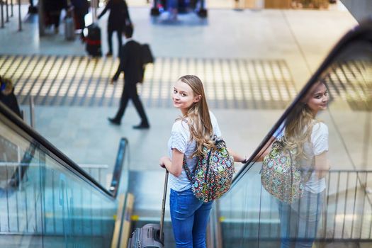 Beautiful young tourist girl with backpack and carry on luggage in international airport, on escalator