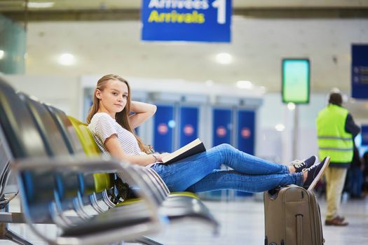 Young traveler with carry on luggage in international airport reading a book while waiting for her flight