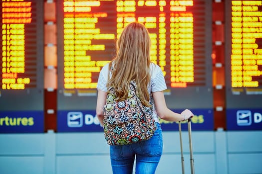 Beautiful young tourist girl with backpack and carry on luggage in international airport, near flight information board