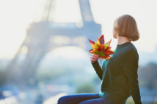 Beautiful young French woman with bunch of colorful autumn leaves near the Eiffel tower in Paris on a fall day