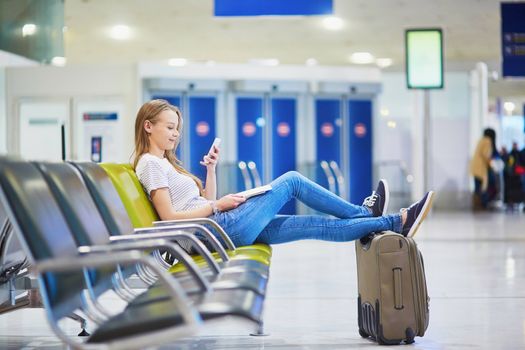 Young traveler with carry on luggage in international airport checking her mobile phone while waiting for her flight