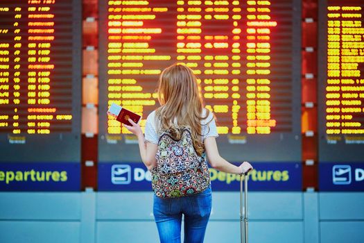 Beautiful young tourist girl with backpack and carry on luggage in international airport, near flight information board