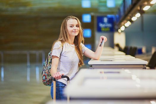 Beautiful young tourist girl with backpack and carry on luggage in international airport at check-in counter, giving her passport to an officer and waiting for her boarding pass