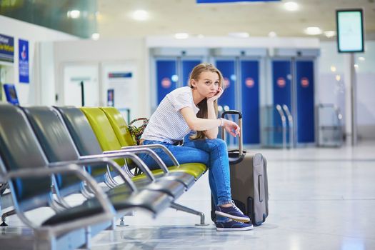 Beautiful young tourist girl with backpack and carry on luggage in international airport, waiting for her flight, looking upset. Delayed or canceled flight concept