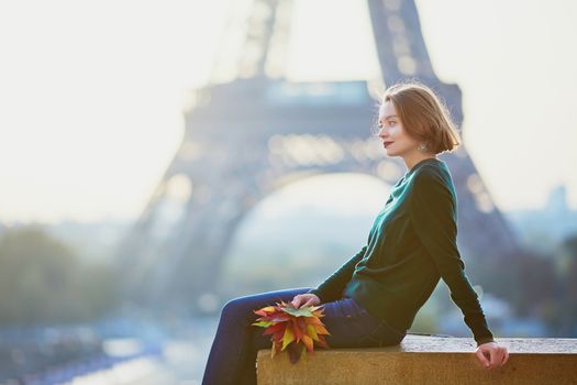 Beautiful young French woman with bunch of colorful autumn leaves near the Eiffel tower in Paris on a fall day