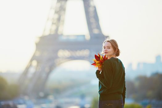 Beautiful young French woman with bunch of colorful autumn leaves near the Eiffel tower in Paris on a fall day