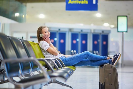 Beautiful young tourist girl with backpack and carry on luggage in international airport, speaking on the phone while waiting for her flight