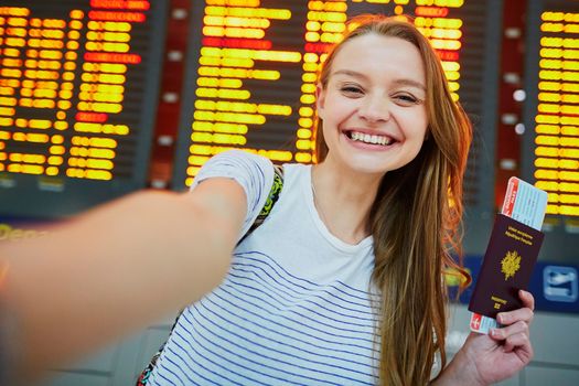Beautiful young tourist girl in international airport, taking funny selfie with passport and boarding pass near flight information board