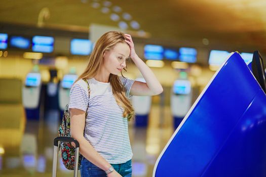 Young tourist girl with backpack and carry on luggage in international airport, doing self check-in, looking upset and worried. Delayed or missed flight concept