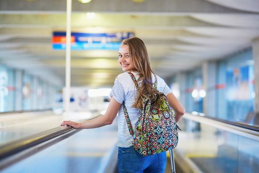 Beautiful young tourist girl with backpack and carry on luggage in international airport, on travelator