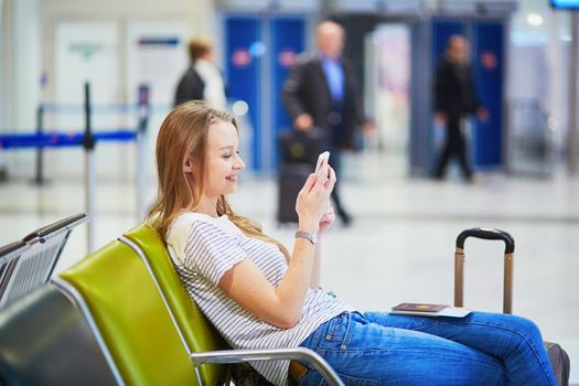 Young traveler with carry on luggage in international airport checking her mobile phone while waiting for her flight