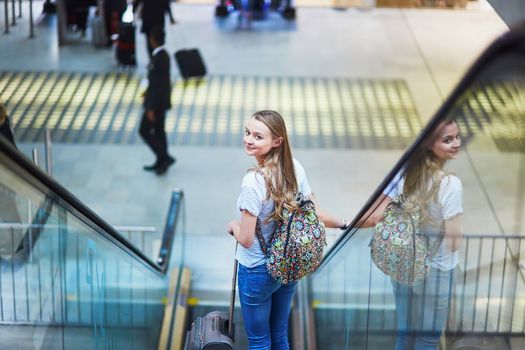 Beautiful young tourist girl with backpack and carry on luggage in international airport, on travelator
