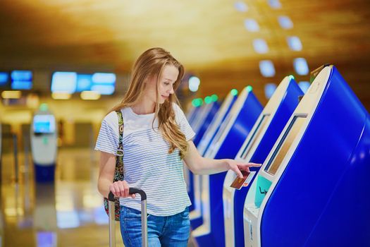 Beautiful young tourist girl with backpack and carry on luggage in international airport, doing self check-in