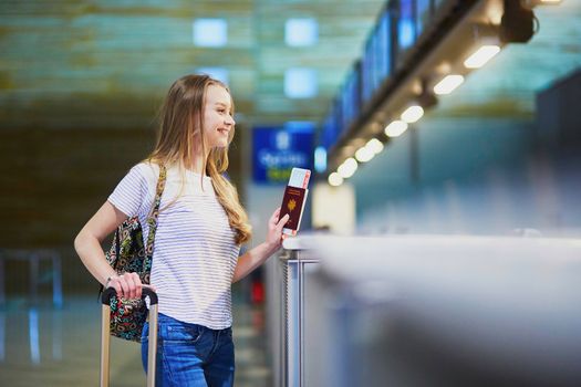 Beautiful young tourist girl with backpack and carry on luggage in international airport at check-in counter, giving her passport to an officer and waiting for her boarding pass