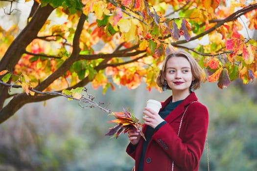 Beautiful young woman with bunch of colorful autumn leaves walking in park on a fall day and drinking coffee to go