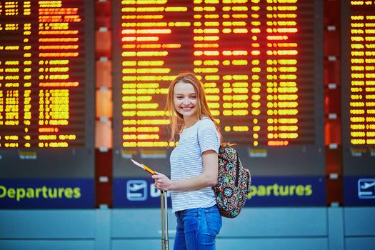 Beautiful young tourist girl with backpack and carry on luggage in international airport, near flight information board